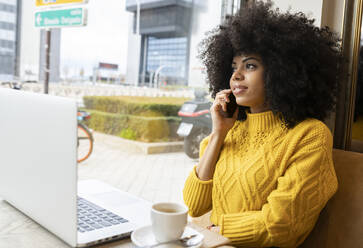 Curly hair woman with laptop talking on mobile phone while sitting at cafe - JCCMF00369