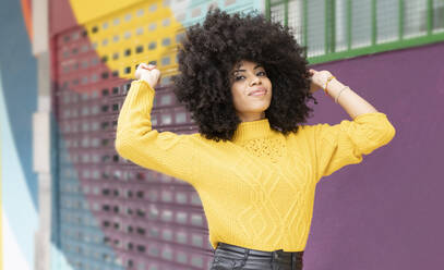 Curly hair woman smiling while standing against multi colored wall - JCCMF00377