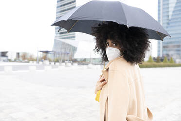 Junge Frau mit Schutzmaske, die einen Regenschirm hält, während sie im Freien steht - JCCMF00398