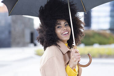 Lächelnde Frau mit Jacke, die einen Regenschirm hält, während sie im Freien steht - JCCMF00404