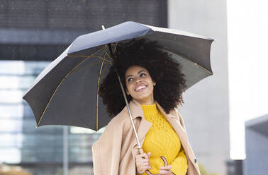 Lächelnde Frau mit Jacke und Regenschirm in der Hand im Regen stehend - JCCMF00406