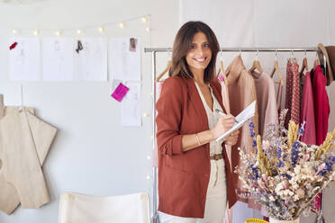 Lächelnde schöne Modedesignerin, die in ihr Tagebuch schreibt, während sie an einem Kleiderständer im Atelier steht - VEGF03407