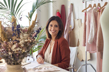 Cheerful female fashion entrepreneur sitting at desk against clothes rack in design studio - VEGF03429