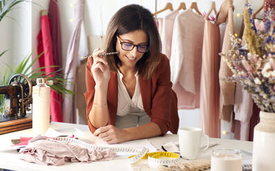 Thoughtful fashion designer smiling while looking at fabric swatch leaning on desk in atelier - VEGF03431