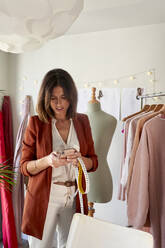 Weibliche Modeunternehmerin, die ihr Smartphone an einem Kleiderständer im Designstudio benutzt - VEGF03444
