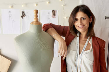 Smiling beautiful fashion designer standing by dressmaker's model at atelier - VEGF03445