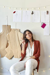 Confident fashion designer sitting with her designs and patterns on white wall at atelier - VEGF03453