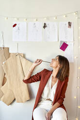 Nachdenkliche Modedesignerin betrachtet ihre Entwürfe und Muster, die an einer weißen Wand im Bekleidungsstudio hängen - VEGF03454
