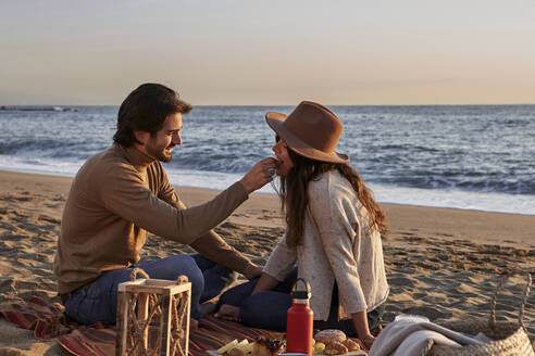 Lächelnder Freund füttert seine Freundin mit Essen, während er am Strand sitzt - VEGF03473