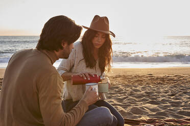 Woman wearing hat pouring coffee in cup while sitting by man on beach - VEGF03475