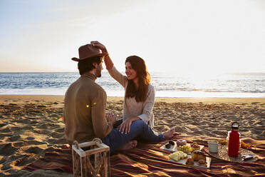 Frau spielt mit Hut und sitzt neben einem Mann am Strand - VEGF03480