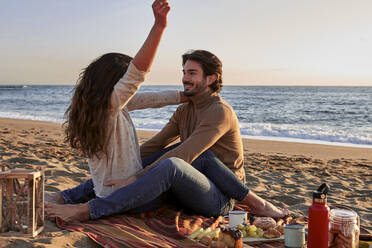 Young woman enjoying while sitting with man on beach during sunrise - VEGF03483