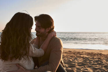 Girlfriend and boyfriend with arm around sitting face to face on beach - VEGF03484