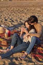 Girlfriend drinking coffee while leaning on boyfriend at beach - VEGF03488