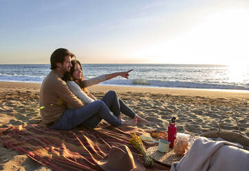 Smiling woman pointing finger toward seawhile sitting with man at beach - VEGF03490