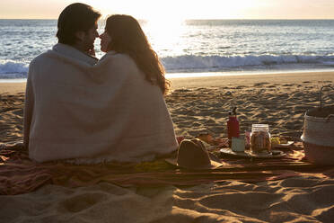 Couple covered in blanket rubbing nose while sitting on beach - VEGF03494