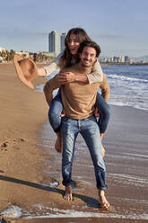 Lächelnder Mann, der eine sorglose Frau huckepack nimmt, während er am Strand steht - VEGF03513