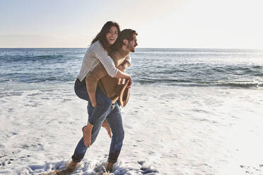 Boyfriend piggybacking smiling girlfriend while walking against sea - VEGF03514