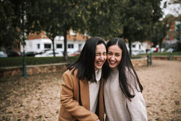 Cheerful sisters standing against trees in park during winter - GRCF00615