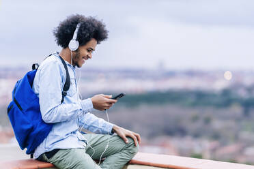 Lächelnder junger Mann hört Musik und benutzt sein Smartphone auf einer Terrasse gegen den Himmel - PGF00317