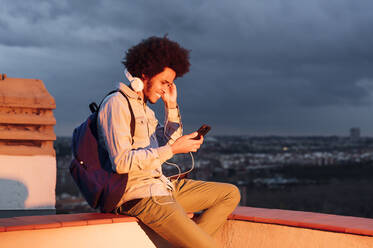 Lächelnder Mann hört Musik und benutzt sein Smartphone auf einer Terrasse bei bewölktem Himmel bei Sonnenuntergang - PGF00327