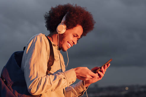 Lächelnder Mann, der Musik hört und sein Smartphone gegen den bewölkten Himmel bei Sonnenuntergang benutzt - PGF00328
