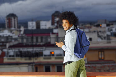 Fröhlicher junger Mann mit blauem Rucksack, der bei Sonnenuntergang vor Gebäuden steht - PGF00333