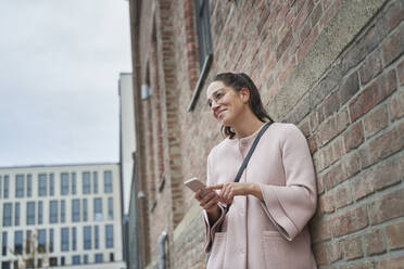 Smiling businesswoman looking away holding smart phone while leaning on brick wall - SDAHF01049