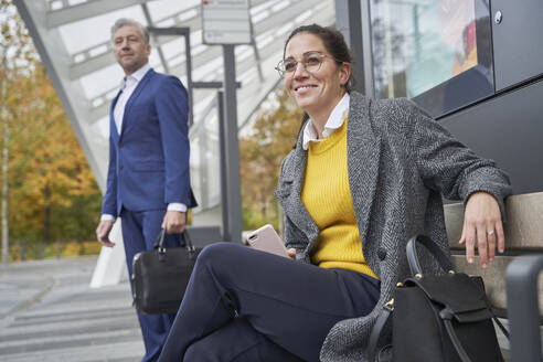 Lächelnde Geschäftsfrau sitzt auf einer Bank, während ein Geschäftsmann an der Bushaltestelle wartet - SDAHF01054