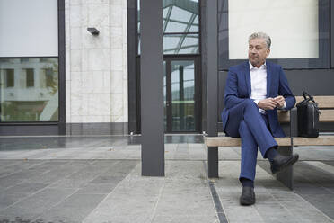 Senior businessman waiting while sitting with legs crossed at bus stop in city - SDAHF01060