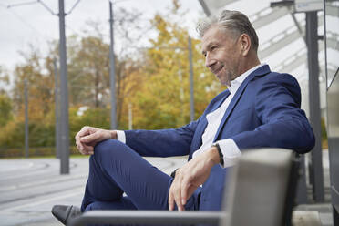 Lächelnde selbstbewusste ältere männliche Fachkraft, die auf einer Bank sitzt und an der Bushaltestelle wartet - SDAHF01068