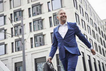 Smiling senior male entrepreneur with bag walking against building in city - SDAHF01078