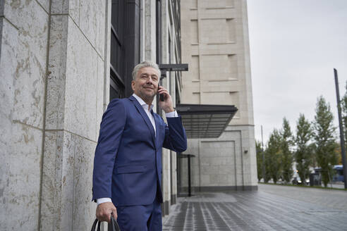 Lächelnder älterer männlicher Unternehmer, der mit seinem Handy telefoniert, während er an einem Gebäude in der Stadt steht - SDAHF01091