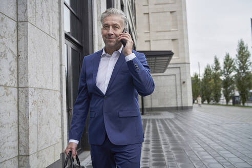 Älterer männlicher Unternehmer, der mit seinem Handy telefoniert, während er an einem Gebäude in der Stadt vorbeigeht - SDAHF01092