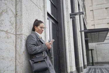 Geschäftsfrau benutzt Smartphone, während sie sich an eine Hauswand lehnt - SDAHF01093
