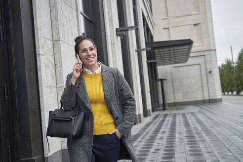 Lächelnde Geschäftsfrau, die mit ihrem Handy telefoniert, während sie in der Stadt spazieren geht - SDAHF01094