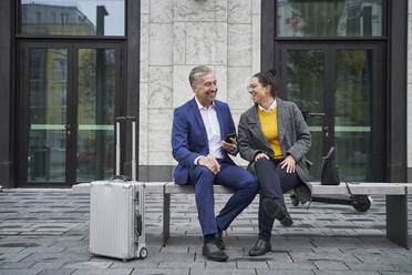 Cheerful male and female professionals talking while sitting on bench against building - SDAHF01097