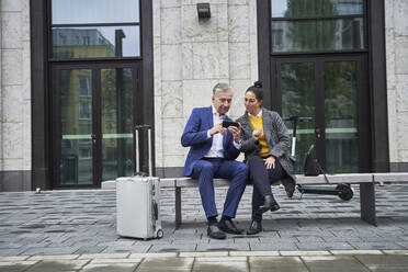 Senior businessman sharing smart phone with female professional while sitting on bench against building - SDAHF01098