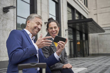 Cheerful male and female entrepreneurs sharing smart phone while sitting against building - SDAHF01099