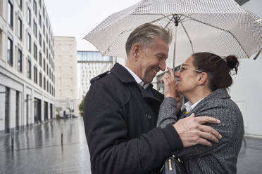 Verspielte Geschäftsfrau mit älterem Geschäftsmann unter Regenschirm in der Stadt während der Regenzeit stehend - SDAHF01104