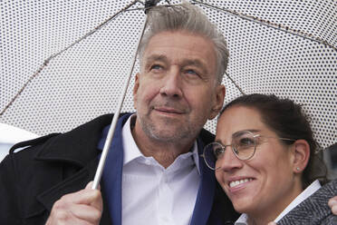 Lächelnde Geschäftsfrau mit älterem Geschäftsmann, der während der Regenzeit unter einem Regenschirm steht - SDAHF01106