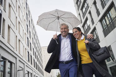 Fröhliche Geschäftsfrau und älterer Geschäftsmann mit Regenschirm beim Laufen in der Stadt während der Regenzeit - SDAHF01108