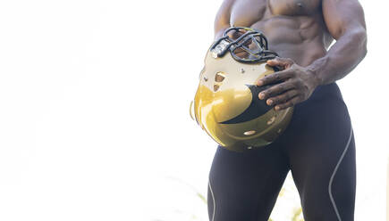 Male athlete holding sports helmet against clear sky on sunny day - JCCMF00440