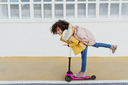 Portrait of cheerful girl with curly hair riding push scooter on road by building - EBBF01968