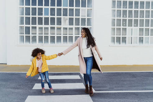 Cheerful mother and daughter holding hands while walking on road against building - EBBF01975