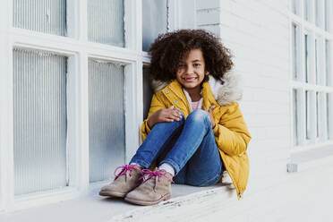 Porträt eines lächelnden Mädchens mit lockigem Haar und gelber Jacke auf dem Fensterbrett sitzend - EBBF01979