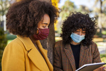 Women wearing protective face mask using digital tablet while leaning on tree at park - MRRF00727