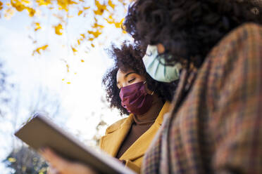 Woman wearing face mask holding digital tablet while standing by friend at park - MRRF00729