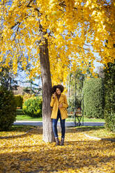 Young woman talking on mobile phone while standing under tree at park - MRRF00731