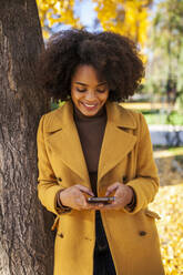 Lockenköpfige Frau lächelt, während sie ein Handy benutzt und sich an einen Baum im Park lehnt - MRRF00733